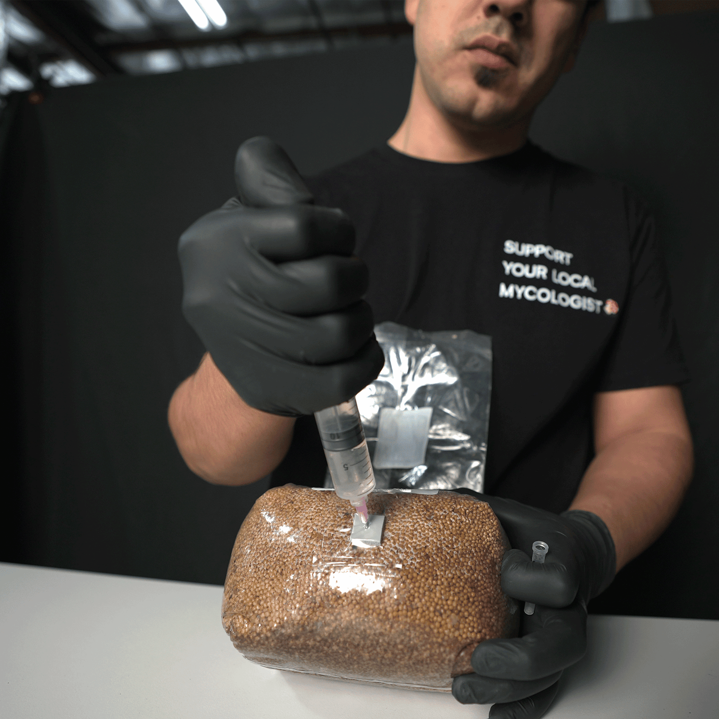 A person in a black t-shirt giving a thumbs up while holding a syringe over the self-healing injection port of the grain spawn bag, ready for inoculation.