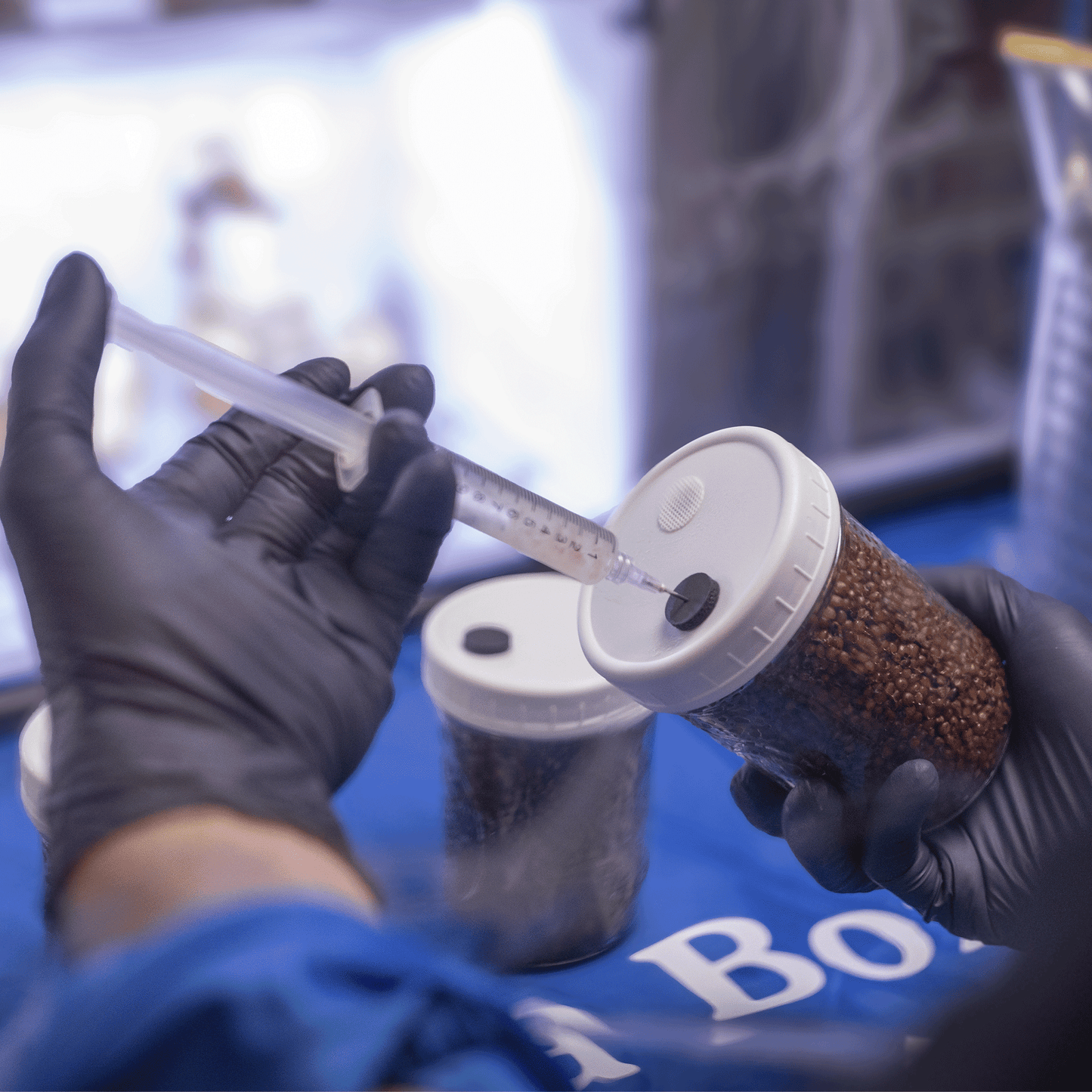 A gloved hand holding a syringe and injecting through a Microppose self-healing injection port on a jar lid in a sterile laboratory environment.